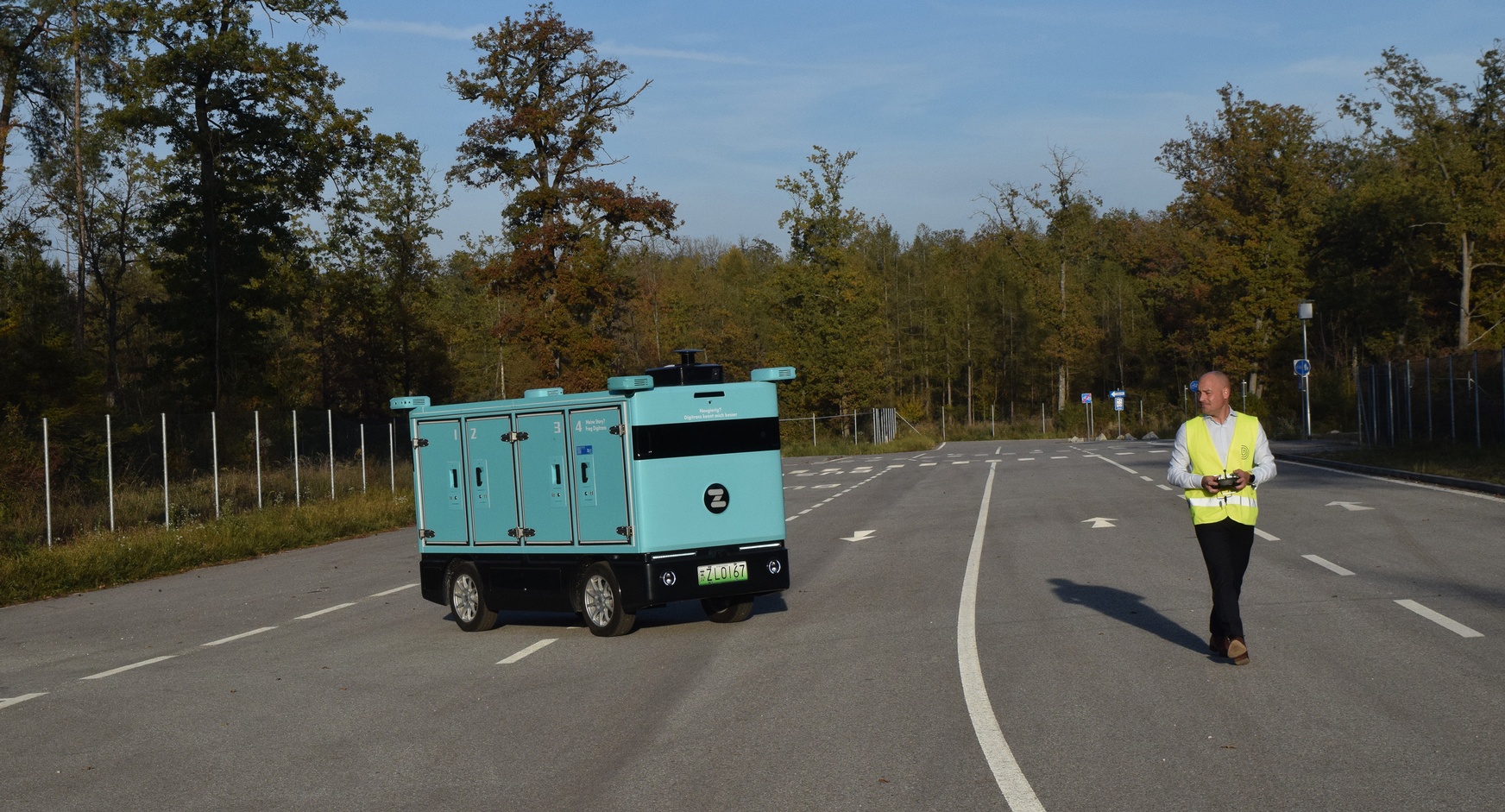 Auf einer mehrspurigen Straße steht ein automatisch fahrendes Fahrzeug mit vier Fächern und vier Rädern. Neben dem Fahrzeug ist eine Person, die das Fahrzeug mit einer Fernbedienung steuert