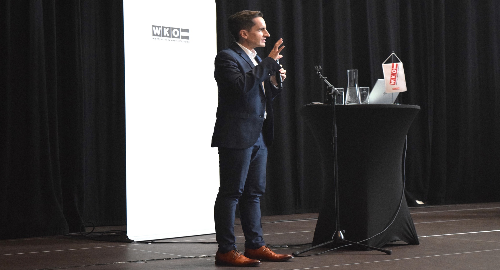 Eine Person steht neben einem Rundtisch, auf dem ein Laptop steht und ein Mikrofon liegt. Die Person spricht in ein Mikrofon. Hinter der Person ist ein weißer Banner, auf dem Wirtschaftskammer Österreich steht