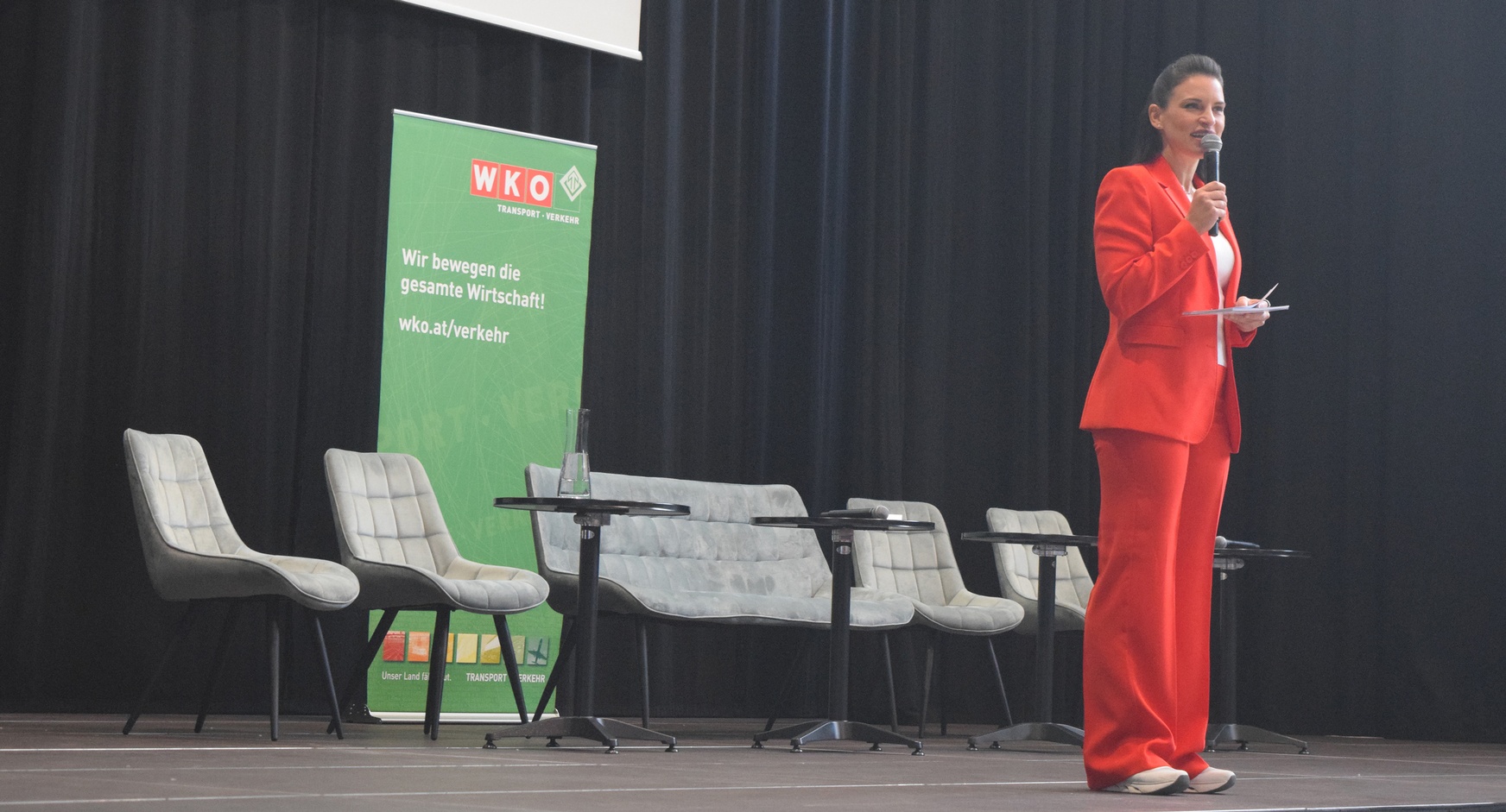 Eine Person in einem roten Anzug und weißem Oberteil steht auf einer Bühne und spricht in ein Mikrofon. Hinter ihr stehen fünf Sessel. Dahinter ist ein grüner Banner, auf dem wir bewegen die gesamte Wirtschaft steht