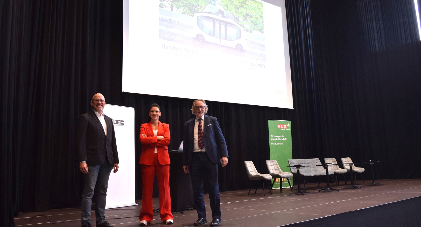 Porträt von drei Personen in Sakkos, die nebeneinander auf einer Bühne stehen. Hinter ihnen ist ein weißer Banner, ein Stehtisch, eine große Leinwand, ein paar Sesseln und ein weiterer Banner, auf dem wir bewegen die gesamte Wirtschaft steht