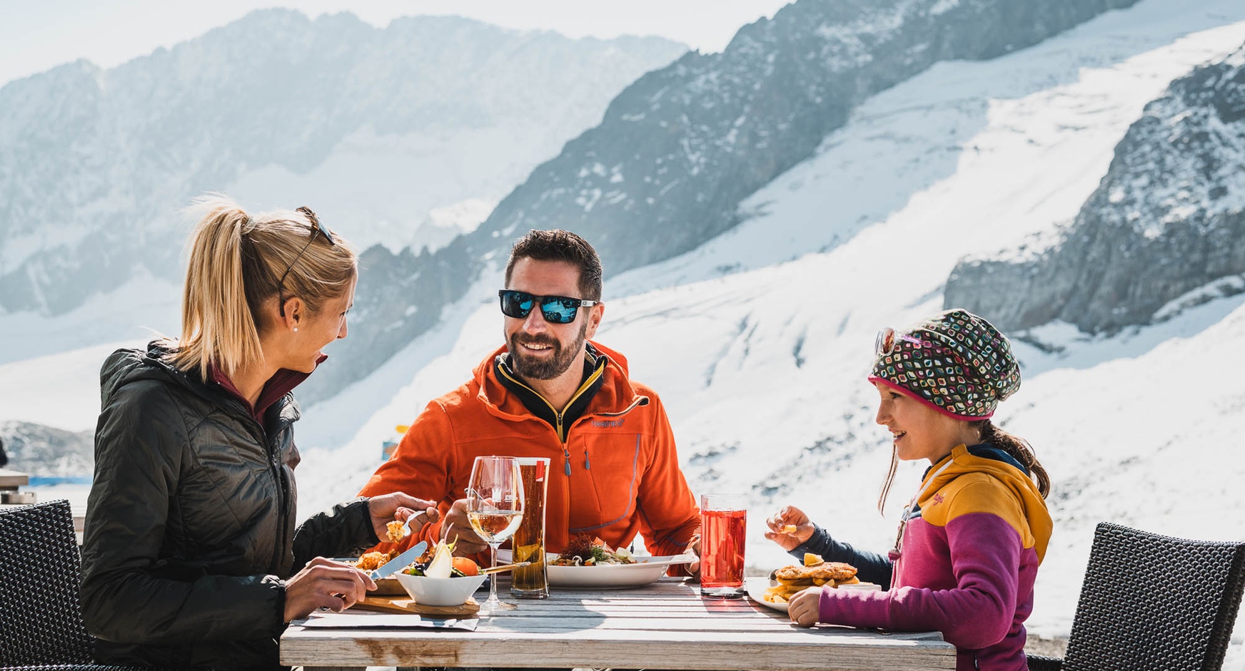 Ein Mann, eine Frau und ein Kind sitzen im Freien an einem Tisch und speisen. Im Hintergrund eine verscheite Berglandschaft.