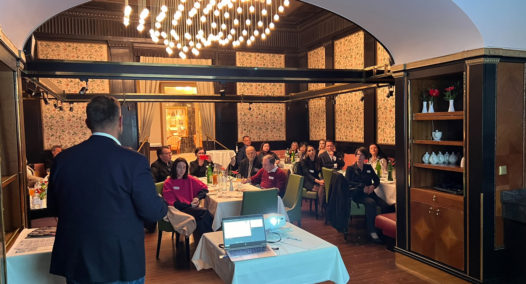 Person in Rückenansicht vor Laptop und auf grünen Stühlen an Tischen sitzendem Publikum sprechend