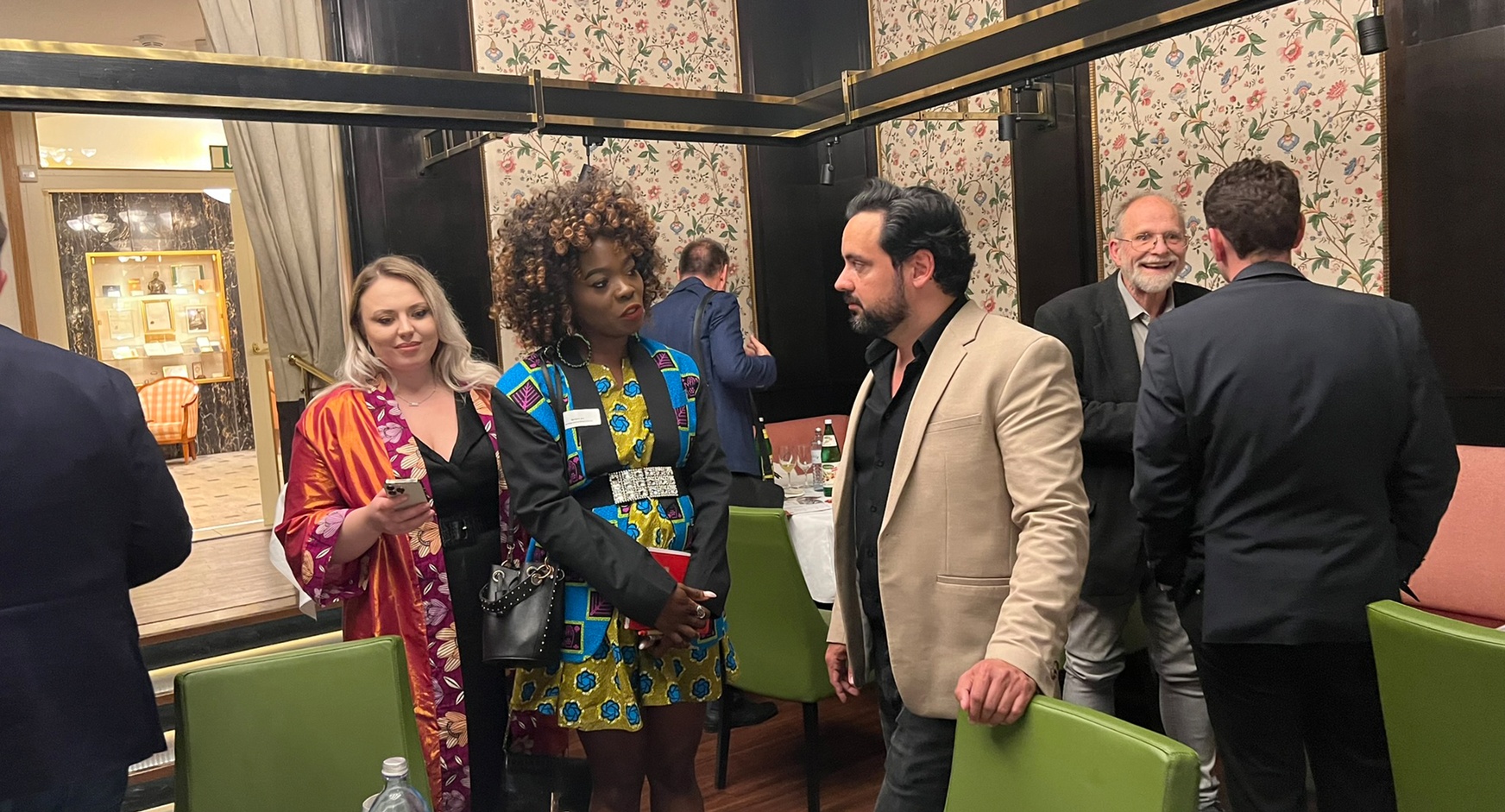 Zwei Personen im Gespräch miteinander, eine Person stützt Hände an grünem Stuhl ab, im Hintergrund floral gemusterte Tapete, Holzvertäfelung und weitere Personen im Gespräch