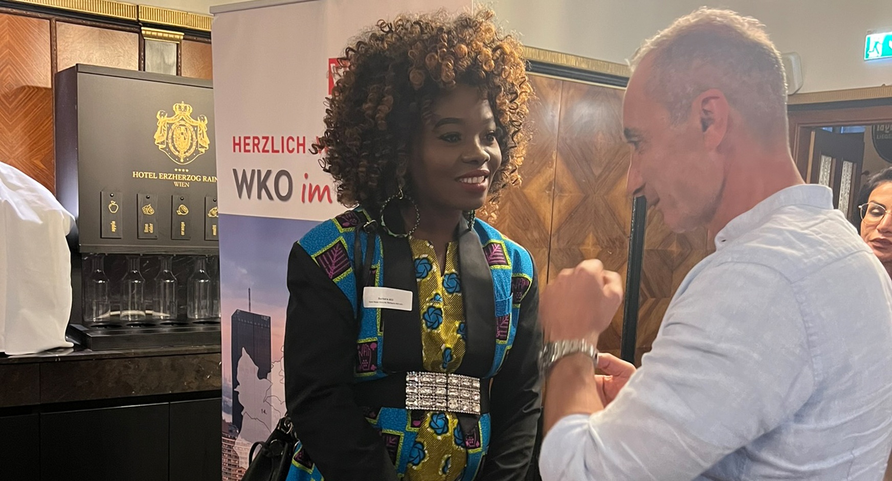 Zwei lächelnde Personen im Gespräch miteinander, im Hintergrund Rollup mit Schriftzug Herzlich WKO im und holzvertäfelte Wand