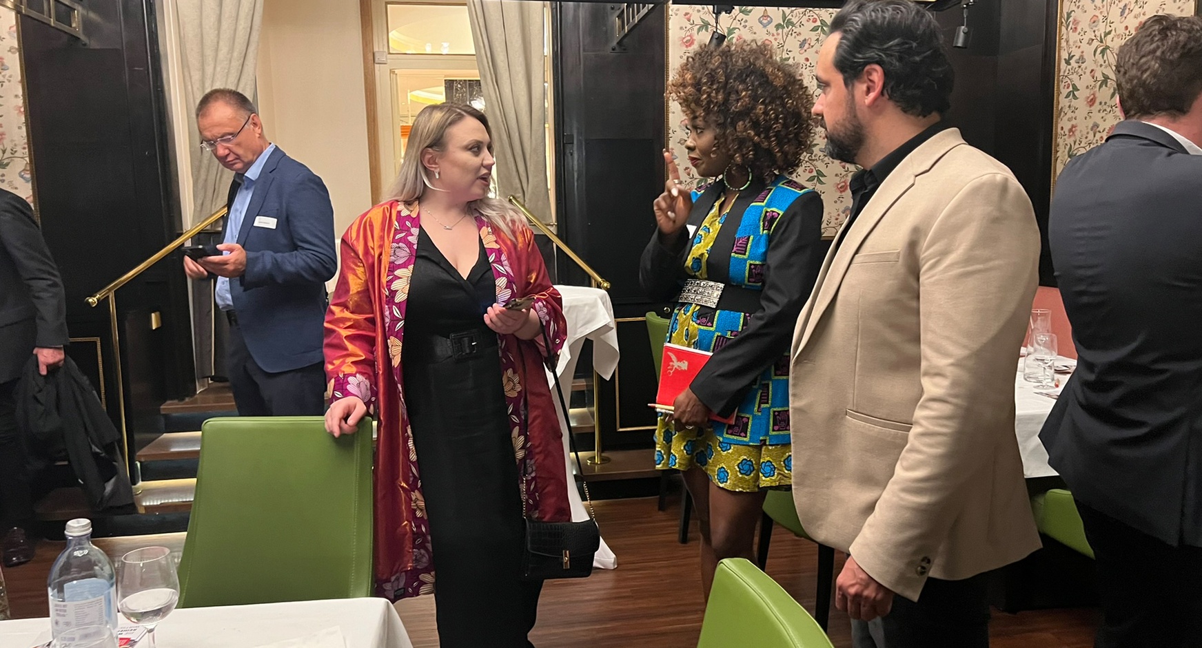 Drei Personen im Gespräch miteinander, eine Person stützt Hände an grünem Stuhl ab, im Hintergrund floral gemusterte Tapete, Holzvertäfelung und weitere Personen im Gespräch