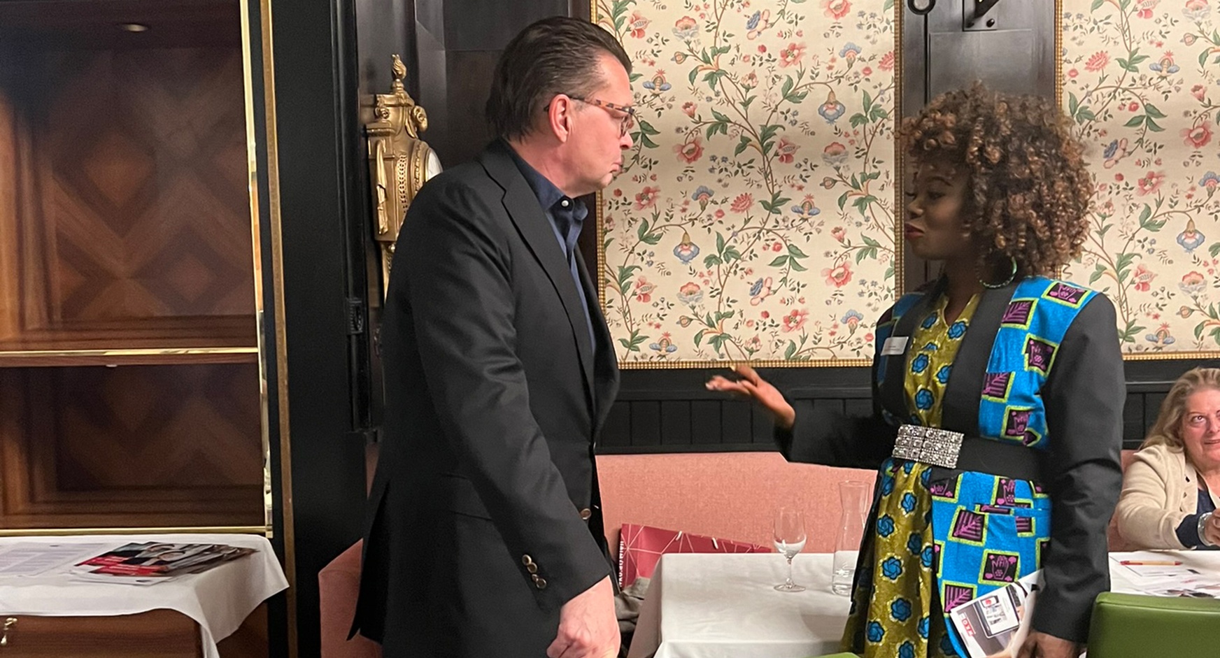 Seitenansicht zweier Personen im Gespräch miteinander, im Hintergrund floral gemusterte Tapete und Holzvertäfelung