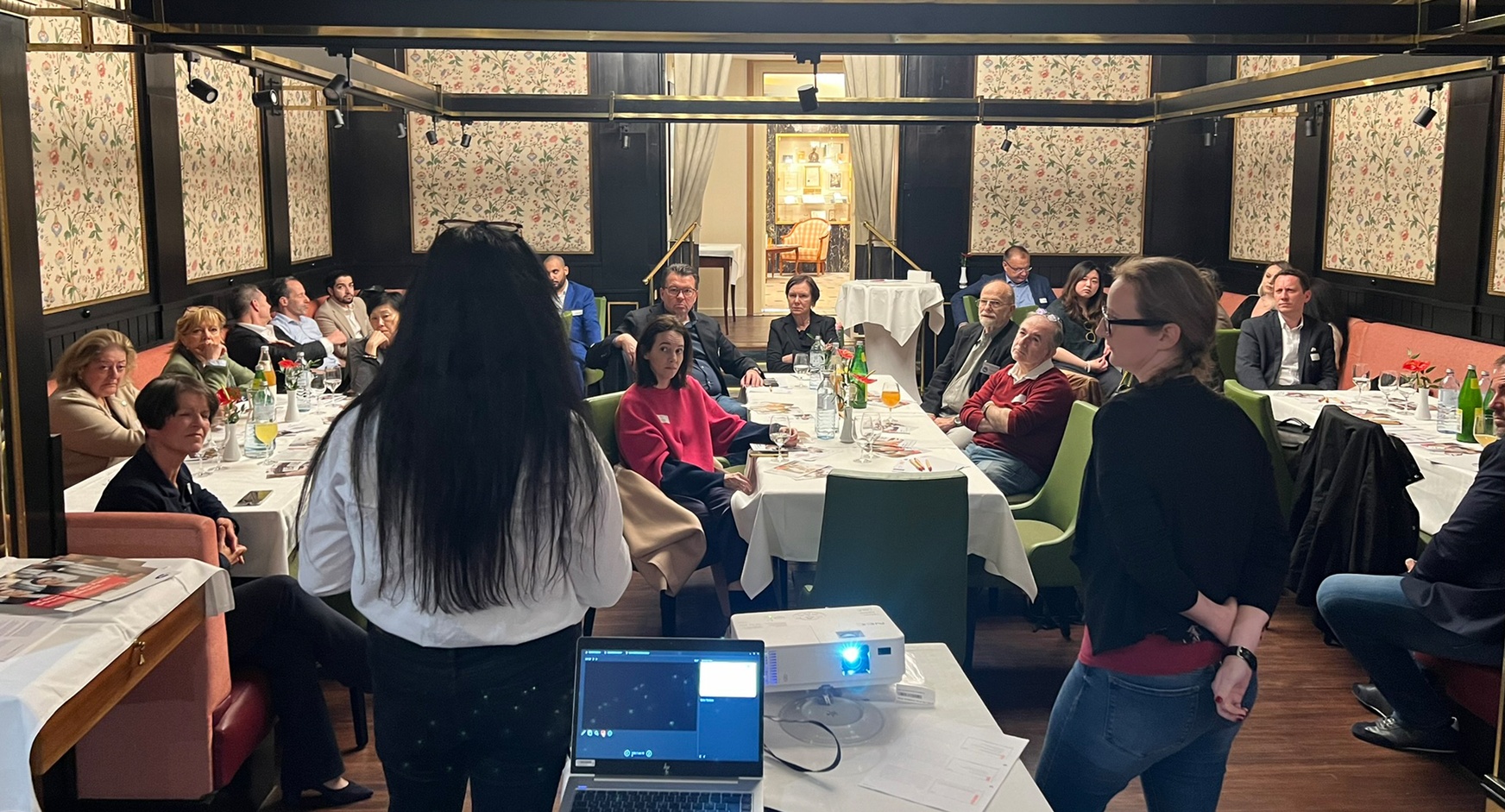 Zwei Personen in Rückenansicht vor Laptop und auf grünen Stühlen an Tischen sitzendem Publikum sprechend