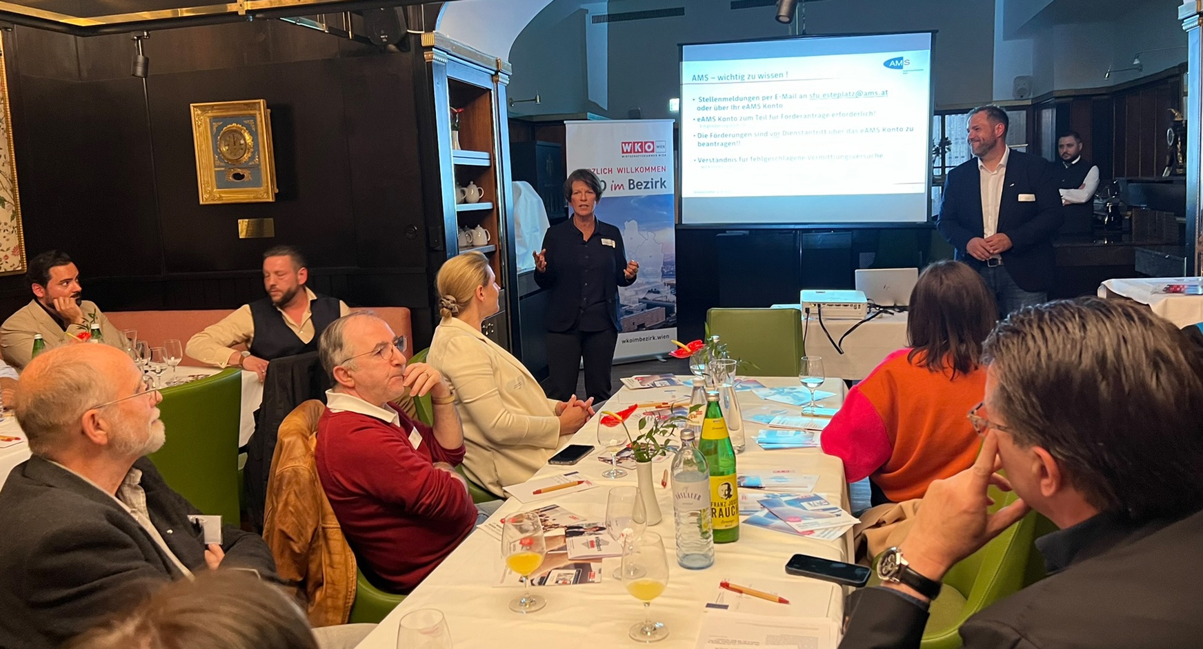 Person vor großer Videowall stehend und vor auf grünen Stühlen an Tischen sitzendem Publikum sprechend