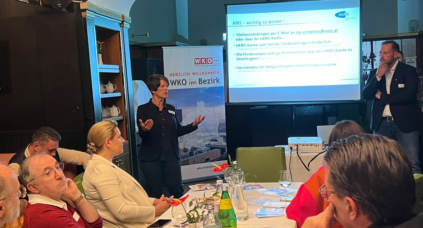 Person vor großer Videowall stehend und vor auf grünen Stühlen an Tischen sitzendem Publikum sprechend