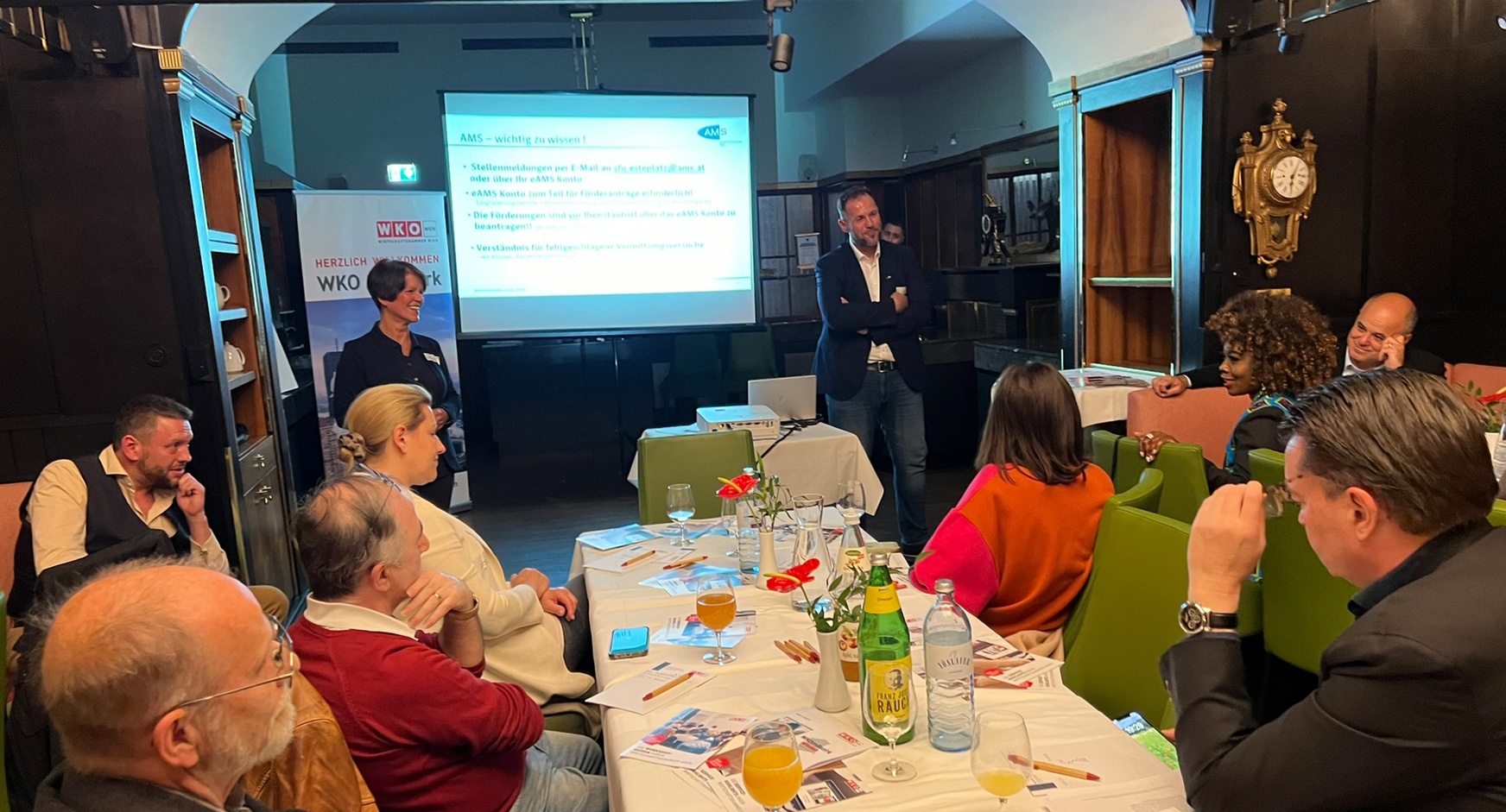 Lächelnde Personen vor großer Videowall stehend und in auf grünen Stühlen an Tischen sitzendem Publikum blickend, eine Person aus dem Publikum spricht