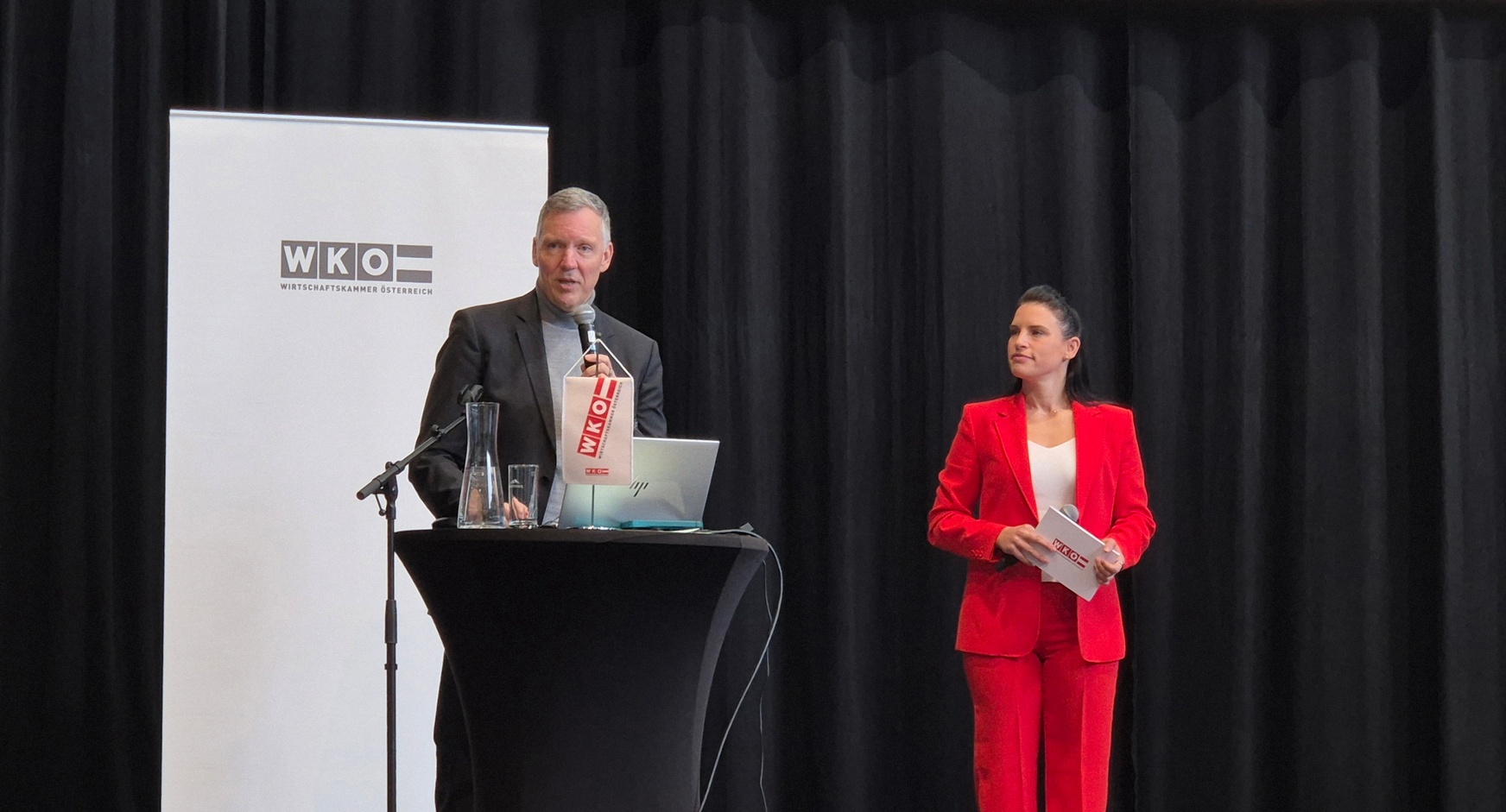 Eine Person steht hinter einem Pult auf einer Bühne und spricht in ein Mikrofon. Rechts neben der Person steht eine weitere Person, die Moderationskarten hält. Hinter der Person hängt eine große Leinwand