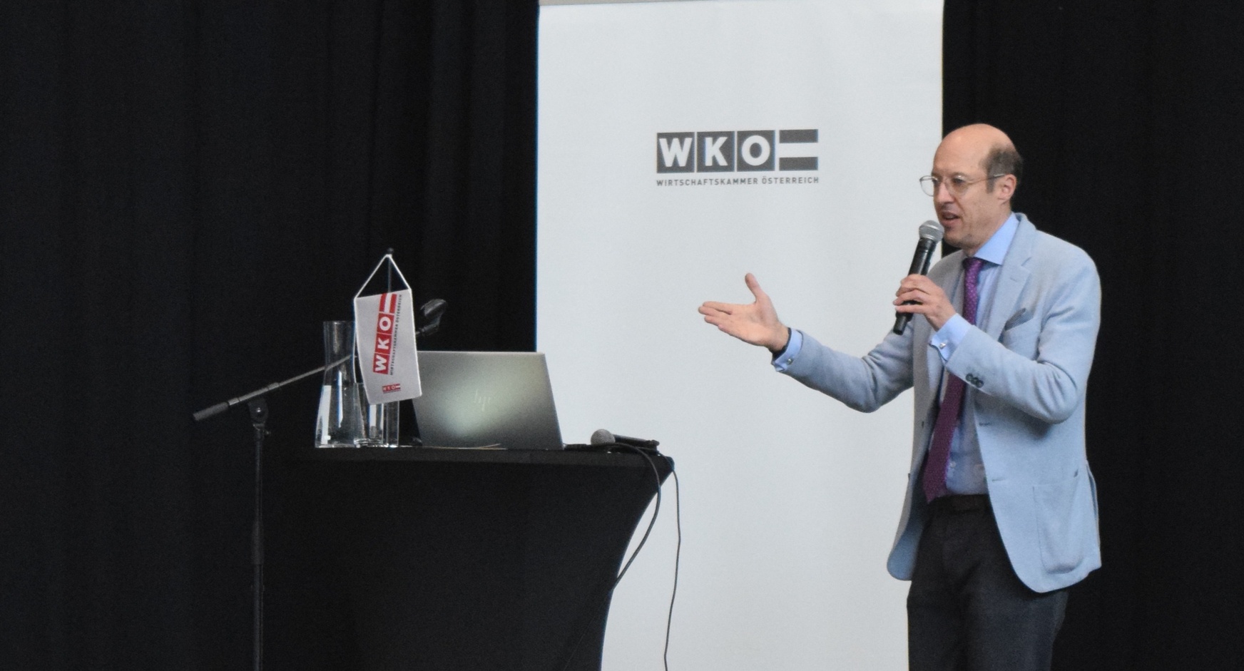 Eine Person steht neben einem Rundtisch, auf dem ein Laptop steht und ein Mikrofon liegt. Die Person spricht in ein Mikrofon. Hinter der Person ist ein weißer Banner, auf dem Wirtschaftskammer Österreich steht