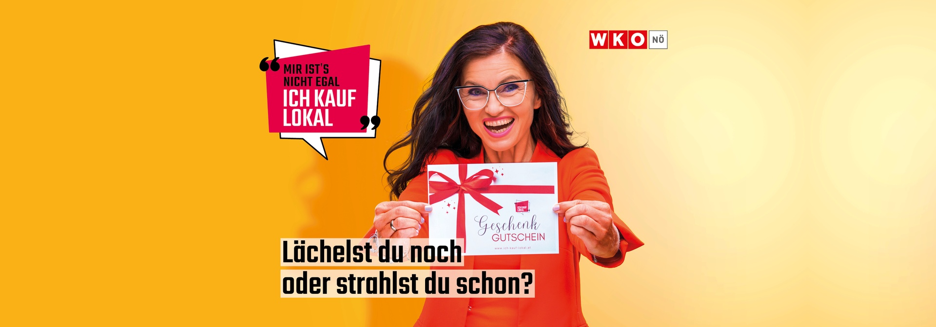 Person in roter Kleidung hält einen Geschenkgutschein mit roter Schleife in beiden Händen. Links oben steht in einer Sprechblase: „Mir ist’s nicht egal – Ich kauf lokal“. Rechts oben das Logo der WKO Niederösterreich. Unten mittig der Text: „Lächelst du noch oder strahlst du schon?“