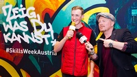 Zwei Personen in Interviewsituation vor Grafiti, beide halten Mikrofone