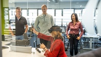 Personen im Büro. Eine davon trägt eine VR-Brille, die anderen schauen dabei zu.