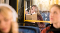 Eine Person sitzt in einem Bus auf einem Sitz und liest in einem Buch. Im Vordergrund verschwommen sind weitere Personen.