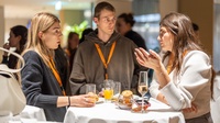Zu sehen sind Personen, die an einem runden Tisch stehen. Auf diesem sind Gläser und Snacks  platziert. 