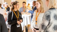 Zu sehen sind Personen, die sich bei einer Veranstaltung unterhalten. Im Hintergrund sind Umrisse anderer Personen und runde Tische zu erkennen. 