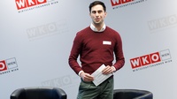Eine Person mit kurzen, dunklen Haaren steht in einem Innenraum vor einer hellgrauen Wand mit mehreren Logos der Wirtschaftskammer Wien. Sie trägt weißes Namensschild auf der Brust und hält mehrere Zettel in den Händen