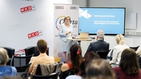 Eine Person mit kurzen, blonden Haaren und Brille steht vor mehreren sitzenden Personen in einem Veranstaltungsraum. Sie trägt ein Namensschild. Auf einem Bildschirm neben ihr steht Kulturwandel im Fokus