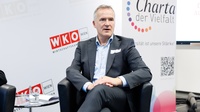 Eine Person mit kurzen, grauen Haaren sitzt in einem schwarzen Ledersessel in einem Veranstaltungsraum. Im Hintergrund befindet sich eine helle Wand mit mehreren rot-weißen Logos und der Aufschrift „WKO Wien – Wirtschaftskammer Wien“