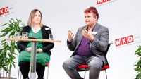Zwei Personen mit Headmikrofonen sitzen auf erhöhten Stühlen neben Stehtisch auf Bühne vor Wand mit rotweißen Logos WKO und Schriftzügen Wirtschaftskammer Wien, eine der Personen spricht und hält Hände vor sich in die Luft