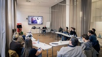 Mehrere Personen sitzen in einem Seminarraum. Vorne sitzt ein Referent und man sieht ein Foto auf einem großen Fernseher.