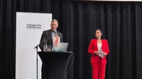 Eine Person steht hinter einem Pult auf einer Bühne und spricht in ein Mikrofon. Rechts neben der Person steht eine weitere Person, die Moderationskarten hält. Hinter der Person hängt eine große Leinwand