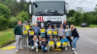 Gruppe von Schulkindern und Begleitpersonen steht vor einem weißen Lkw mit „stl logistics“-Logo; mehrere Kinder halten gelbe Warnmaterialien mit der Aufschrift „LKW FRIENDS“; Aufnahme im Freien auf einem Parkplatz.