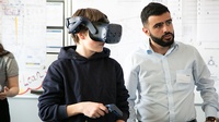Person mit VR-Brille und Kontroller neben weiterer Person stehend