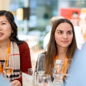 Zu sehen sind Personen, die an einem Tisch stehen, auf dem auch Gläser mit Sekt platziert sind.