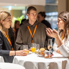 Zu sehen sind Personen, die an einem runden Tisch stehen. Auf diesem sind Gläser und Snacks  platziert. 