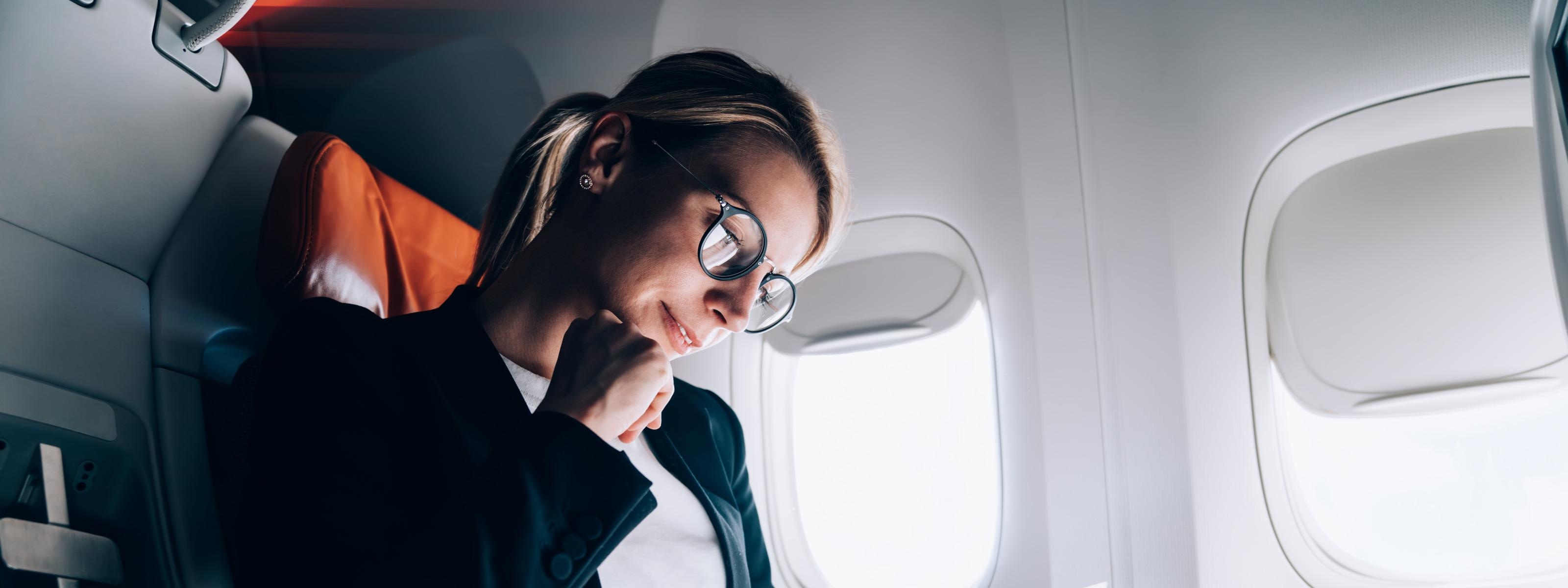 Person mit Brillen sitzt in Flugzeug und blickt auf Dokumente, eine Hand ans Kinn gestützt