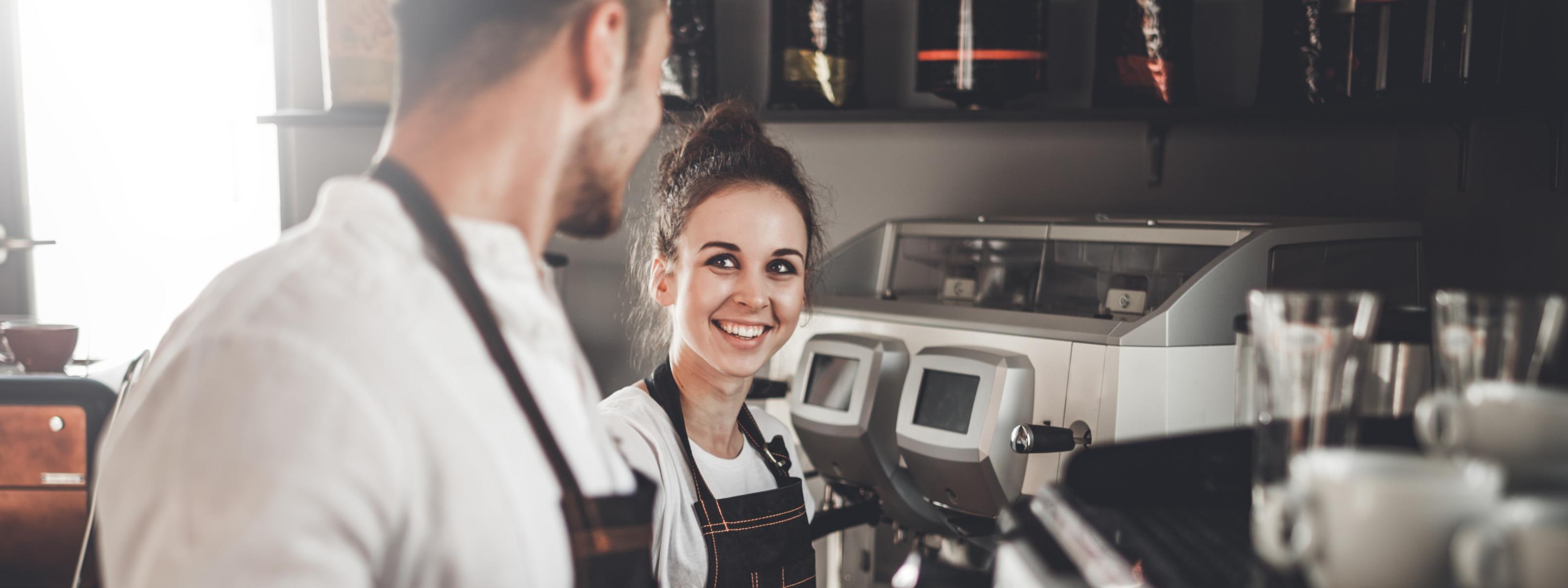 Person lächelt andere Person an, beide tragen Schürzen und stehen vor Kaffemaschine