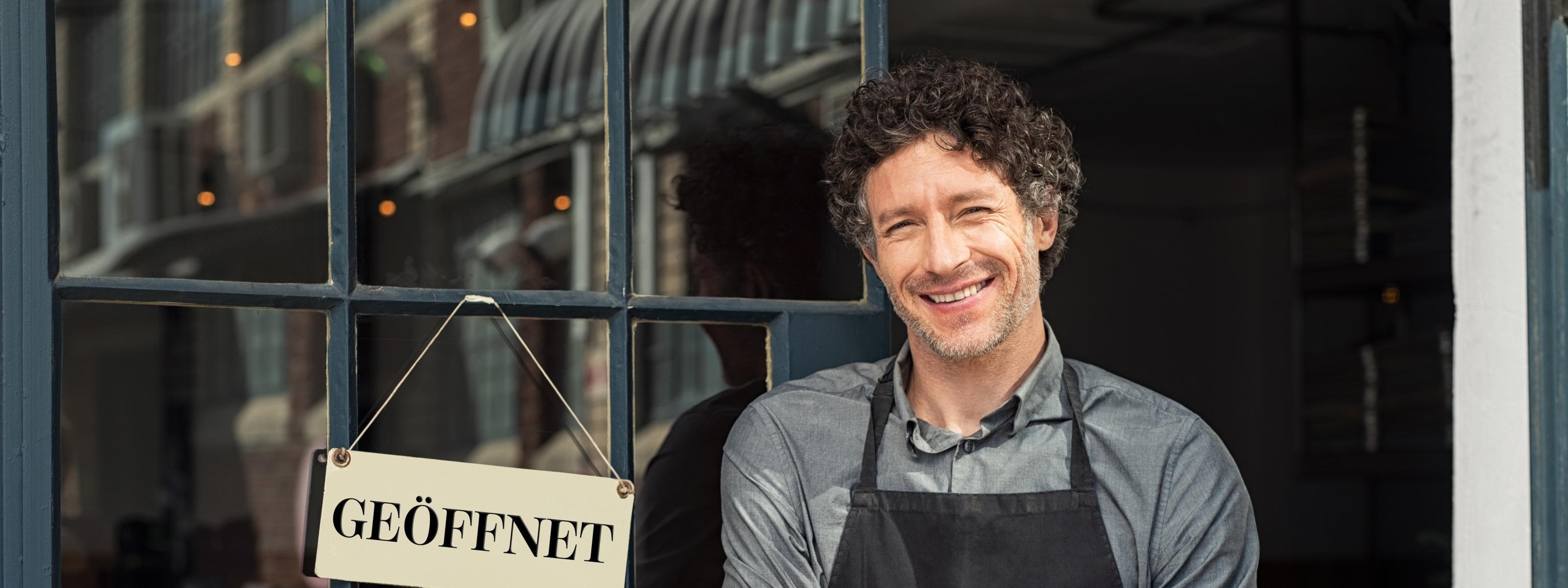 Eine Person steht neben der Lokaltür bei einem Schild GEÖFFNET und lächtelt in die Kamera.