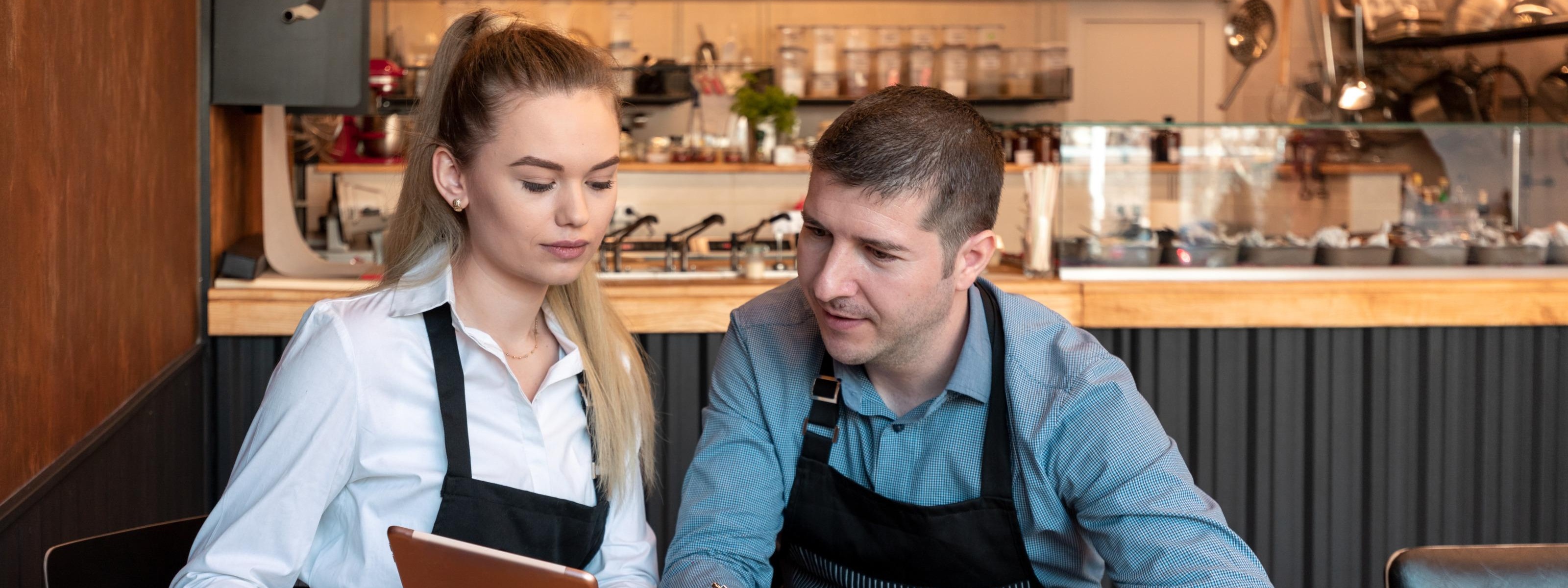Zwei Personen in Arbeitskleidung sitzen an einem Tisch in einem Restaurant und blicken auf ein Tablet während mit einem Taschenrechner gerechnet wird und mit einem Stift Notizen gemacht werden