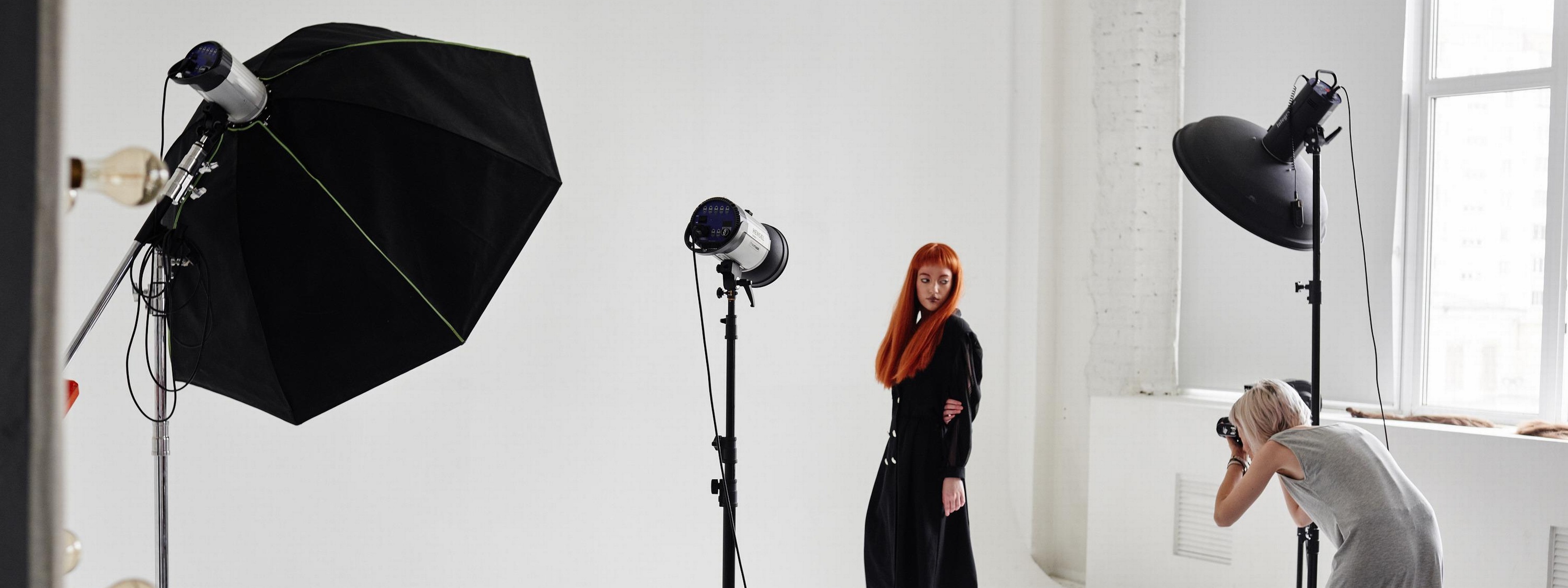 Person in dunklem Kleid und mit langen roten Haaren posiert vor weißem Hintergrund für andere Person, die Fotos mit Kamera macht, ringsum Studioblitze