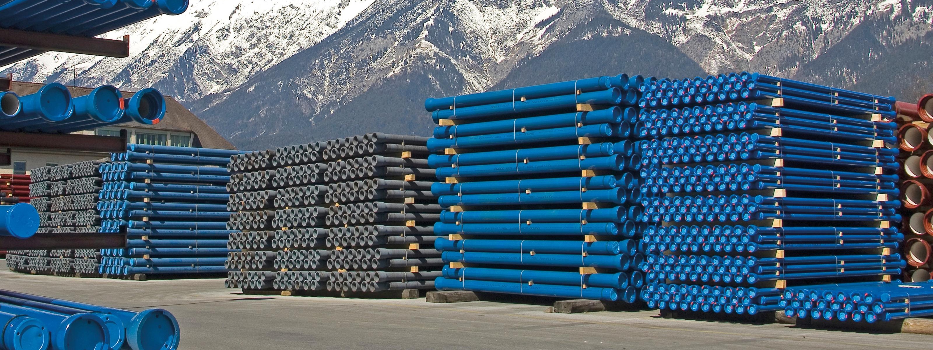 Lagerplatz mit gestapelten blauen Rohren, im Hintergrund beschneite Bergkette und blauer Himmel