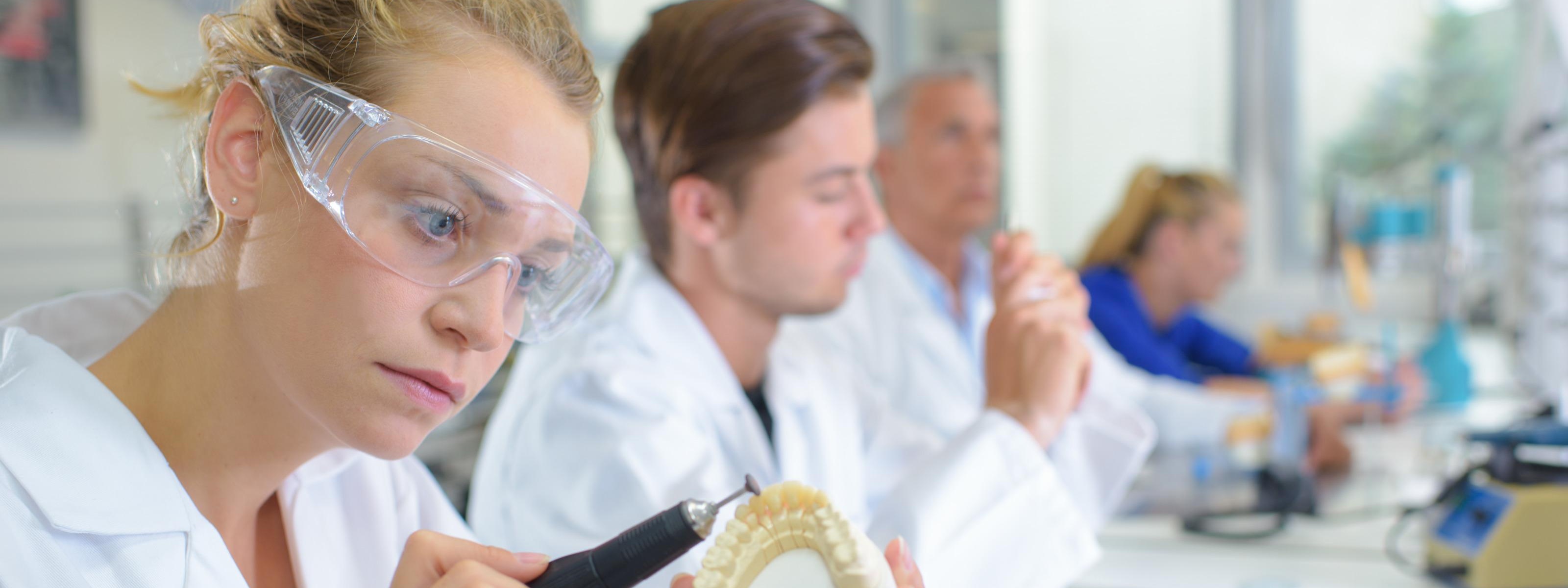Person mit Schutzbrille und weißer Arbeitskleidung arbeitet an einer Zahnprothese mit einem elektrischen Schleifgerät, im Hintergrund sitzen weitere Personen an Arbeitsplätzen