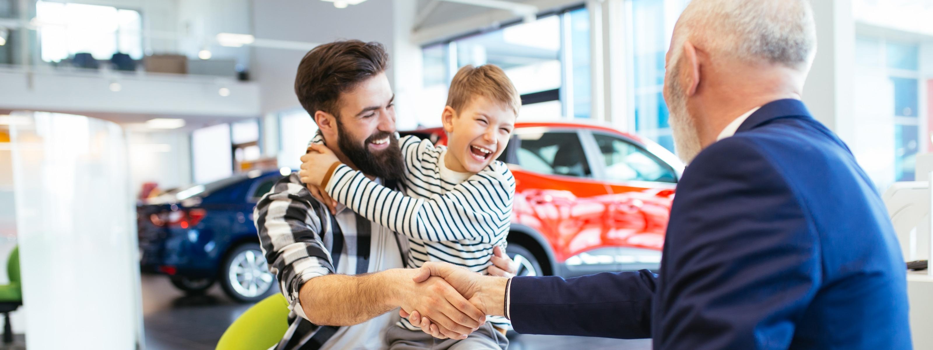 Person mit lachendem Kind am Schoß schüttelt weiterer Person am Tisch sitzend die Hand, im Hintergrund Autos