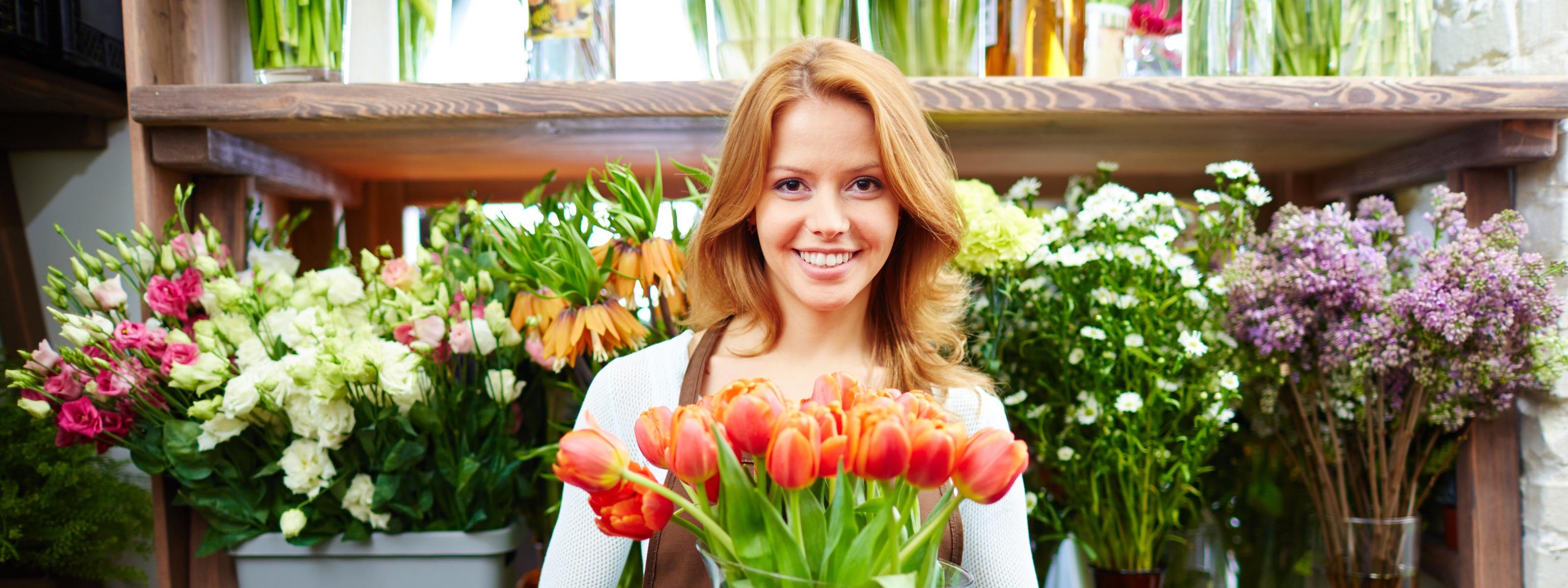 Lächelnde Person mit Schürze hält Vase mit orangen Tulpen in Händen vor Regal mit verschiedenen Schnittblumen stehend