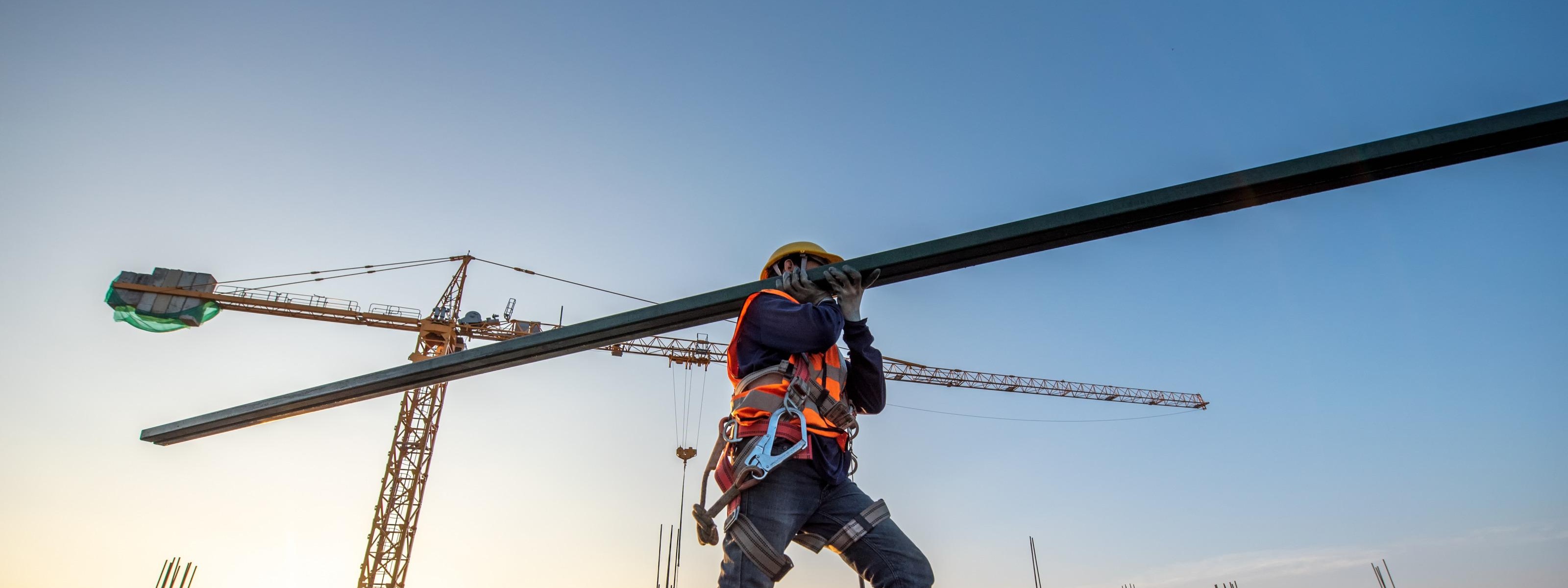 Person ich Schutzkleidung trägt großen Metallbalken über Baugrund, im Hintergrund Kran im Sonnenuntergang