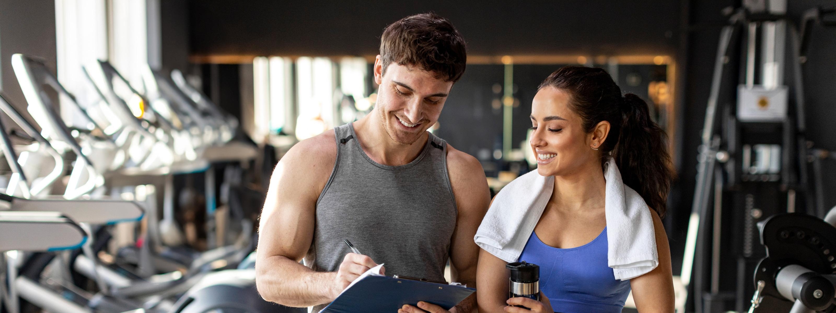 Zwei Personen in Sportkleidung stehen nebeneinander. Die linke Person notiert etwas auf einem Klemmbrett, auf das beide blicken. Die rechte Person hat ein Handtuch um den Hals. Im Hintergrund stehen verschiedene Sportgeräte