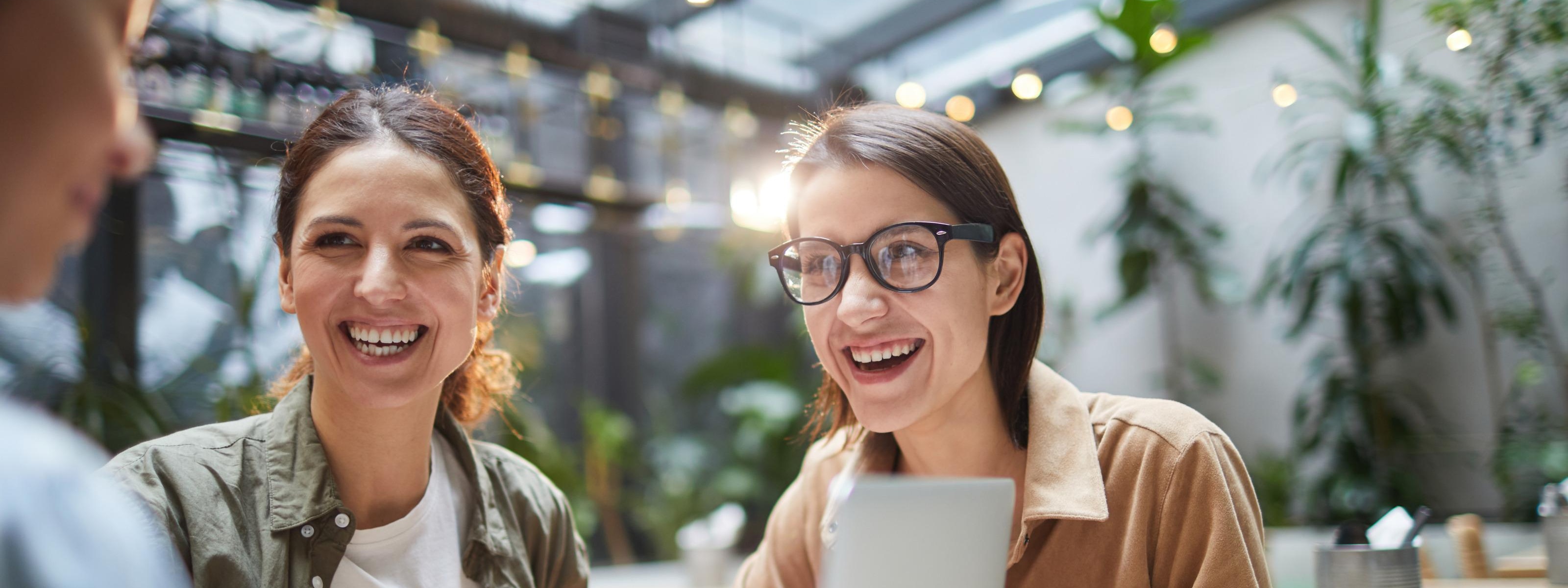 Zwei lächelnde Personen sitzen am Tisch mit weiterer Person vor Laptop, Kaffeetassen am Tisch, im Hintergrund Lichterketten und Pflanzen