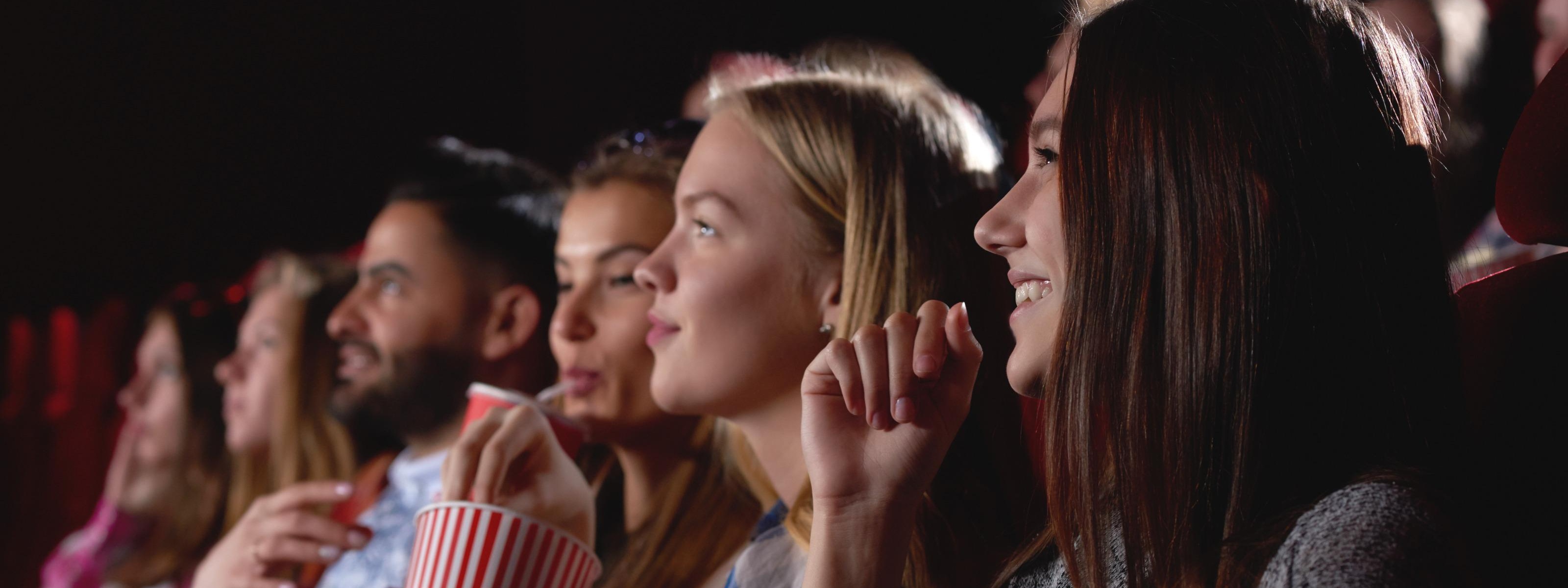 Mehrere Personen nebeneinander in Kinosaal sitzend schauen Richtung Leinwand und halten Getränke und Popcorn in Händen