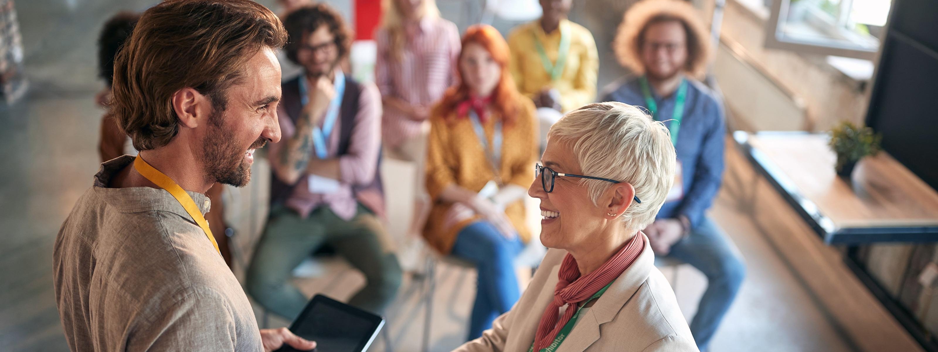 Personen geben sich stehend die Hand während weitere Personen im Hintergrund sitzen und freudig zusehen 