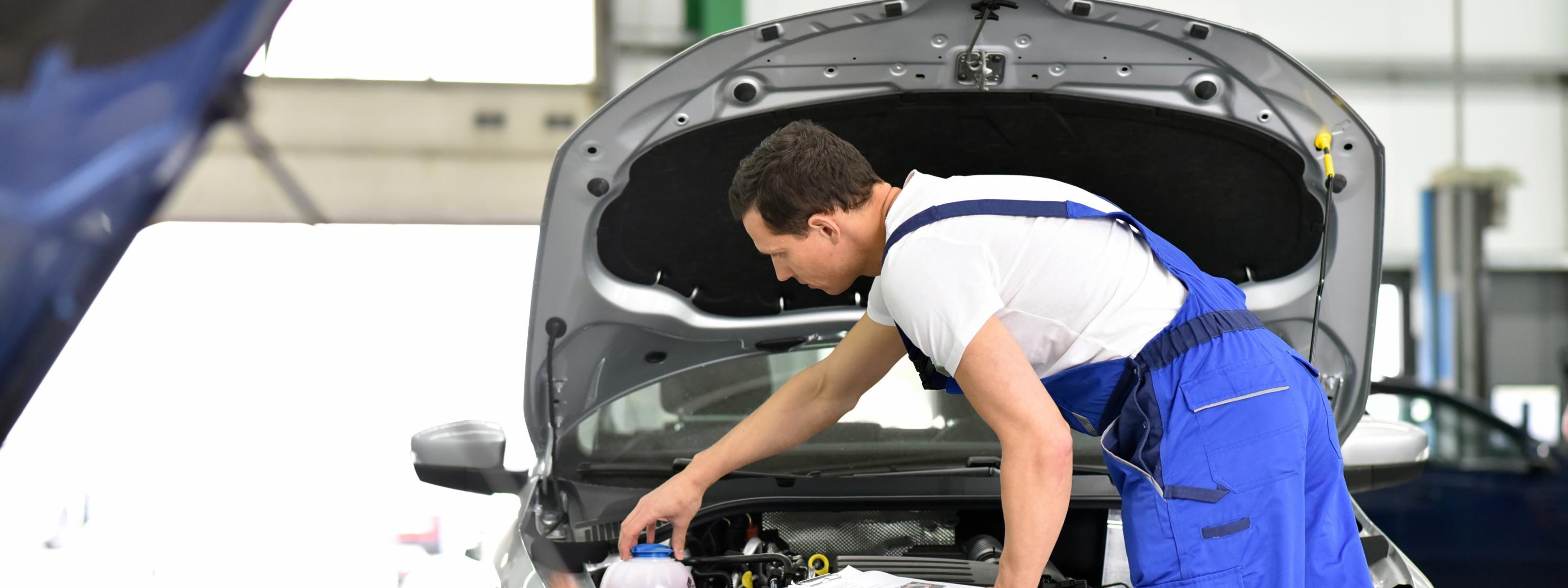 Person mit blauer Arbeitslatzhose beugt sich über Motor eines Autos mit aufgeklappter Motorhaube und schraubt an Tank