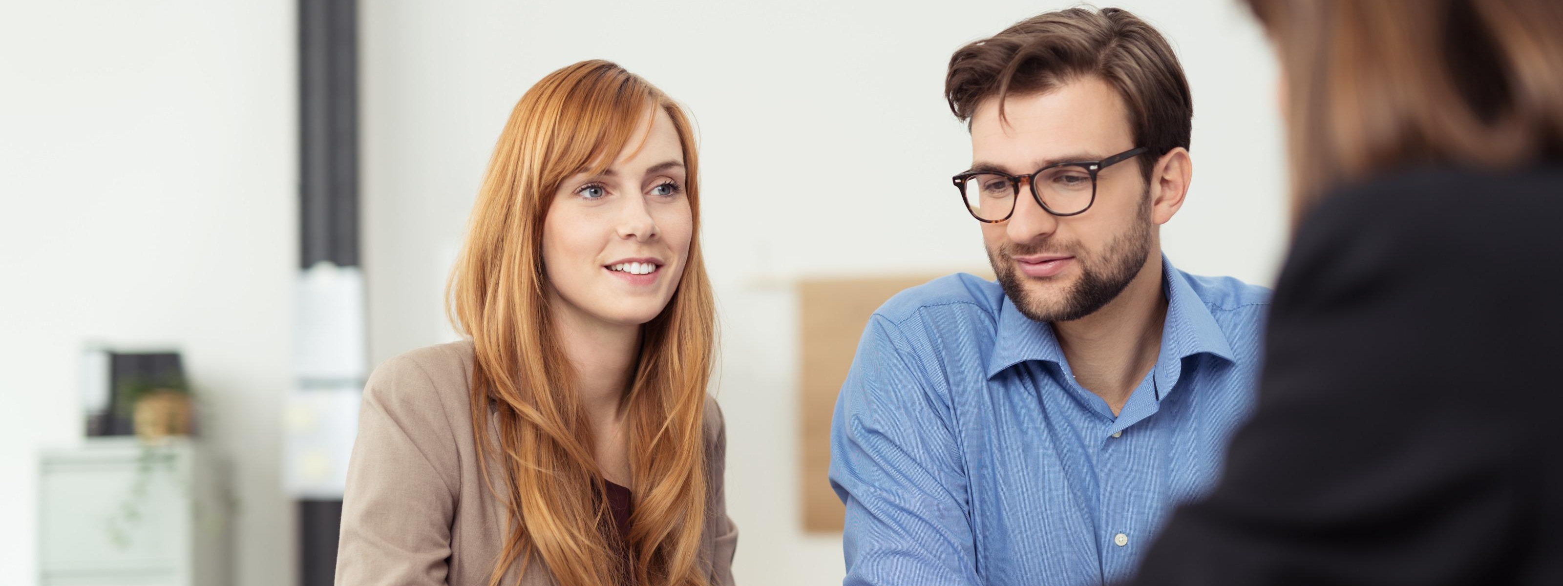 Zwei Personen sitzen an Tisch, eine Person lächelt sanft, die andere trägt Brillen, im Vordergrund verschwommen weitere Person