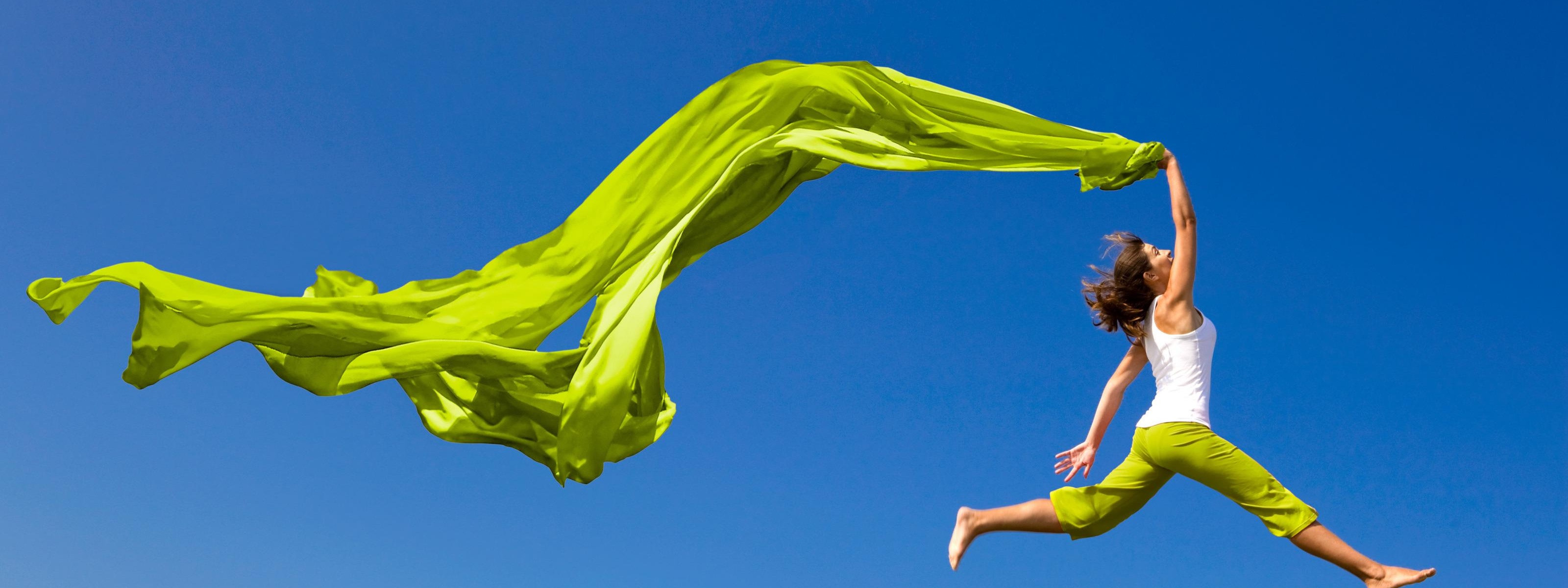 Person in grüner Hose mit grünem großem Tuch kopfüber in der Hand springt über satte Wiese, im Hintergrund blauer Himmel