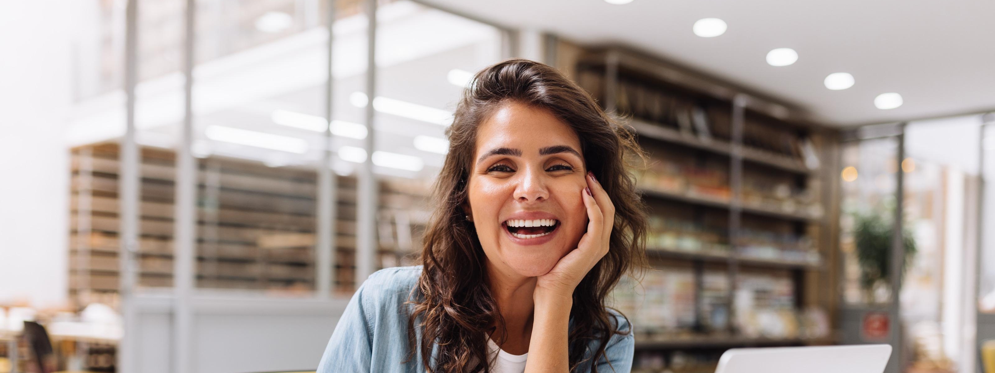 Lachende Person vor Laptop in Großraumbüro sitzend hält in einer Hand ein Smartphone, die andere Hand den Kopf stützend, ringsum Kartonagen